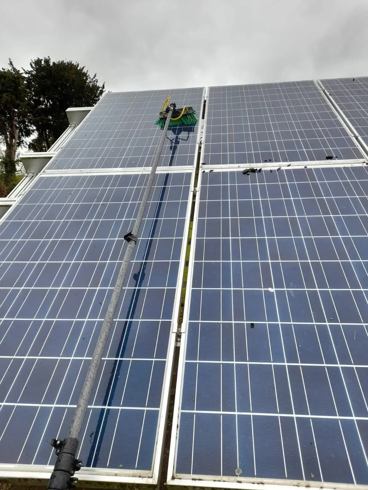 Solar panel array being cleaned with a telescopic brush, with dirt and debris visible on the panels